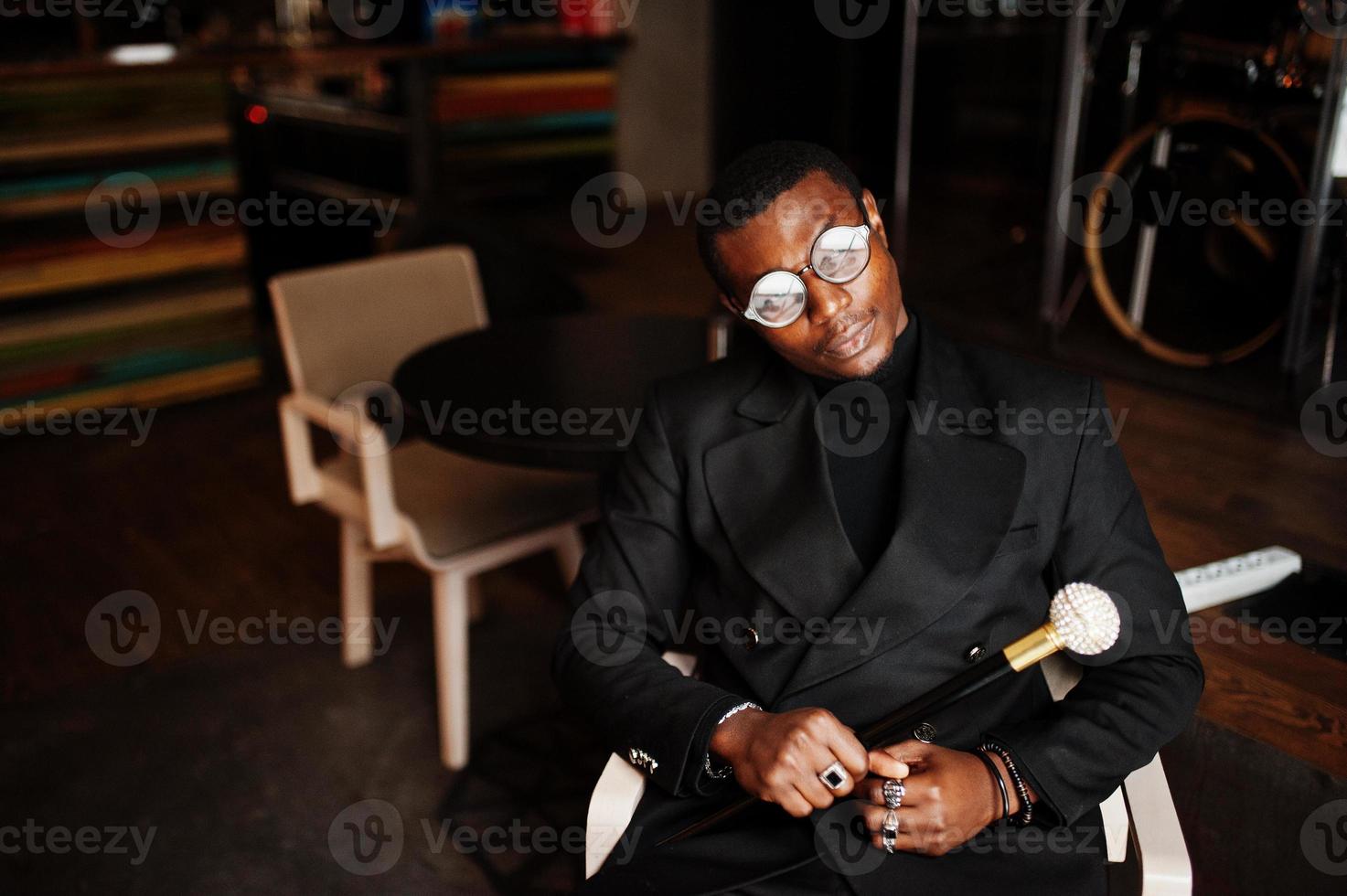 élégant gentleman afro-américain dans une élégante veste noire et des lunettes, tenant un bâton de marche rétro comme un flacon de canne ou une canne basculante avec une poignée en boule de diamant doré. riche homme afro à la mode. photo