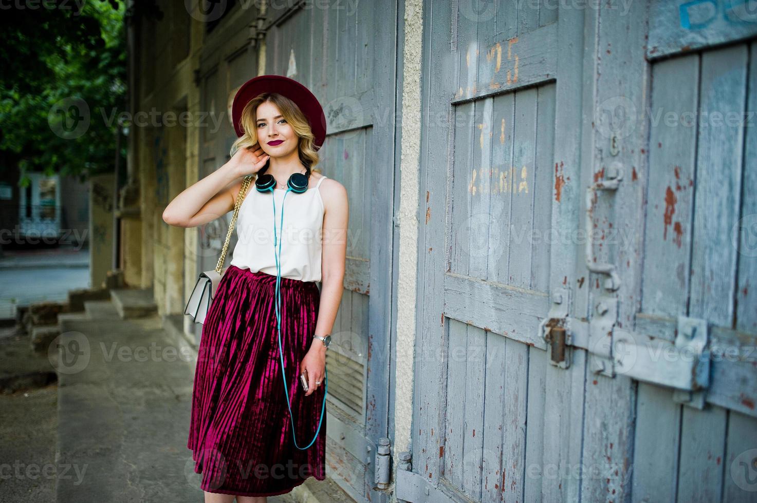 fille modèle blonde à la mode et belle en élégante jupe en velours rouge, chemisier blanc et chapeau, posée avec téléphone et écouteurs. photo