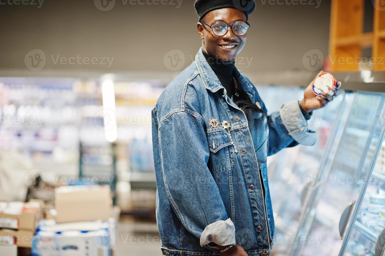 élégant homme afro-américain décontracté à la veste en jean et au béret noir tenant un panier, debout près d'un réfrigérateur gastronomique et faisant ses courses au supermarché. photo