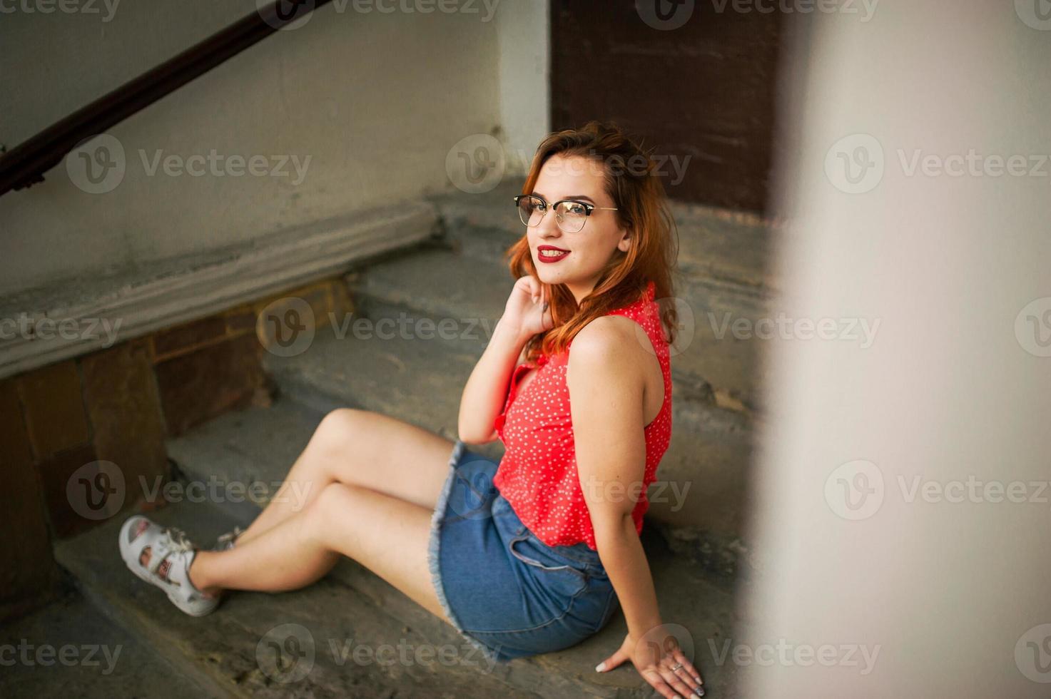 jolie femme rousse à lunettes, porter sur un chemisier rouge et une jupe en jean posant. photo