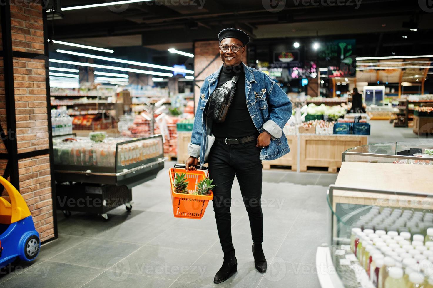 homme afro-américain décontracté et élégant à la veste en jean et au béret noir tenant un panier avec des ananas dans la section bio des fruits du supermarché. photo