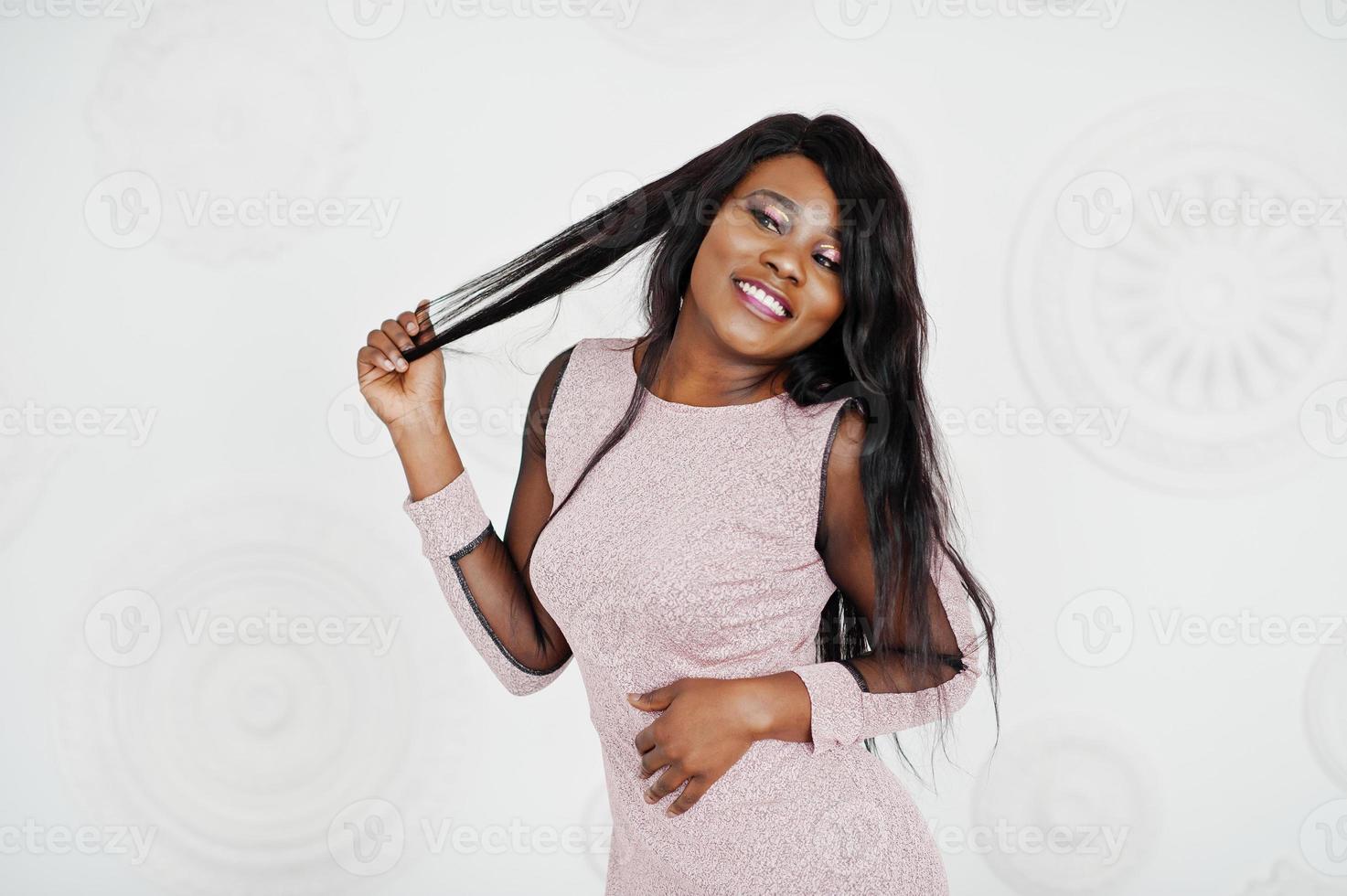 femme modèle afro-américaine à la mode en robe de soirée brillante rose posée contre un mur décoratif blanc. photo