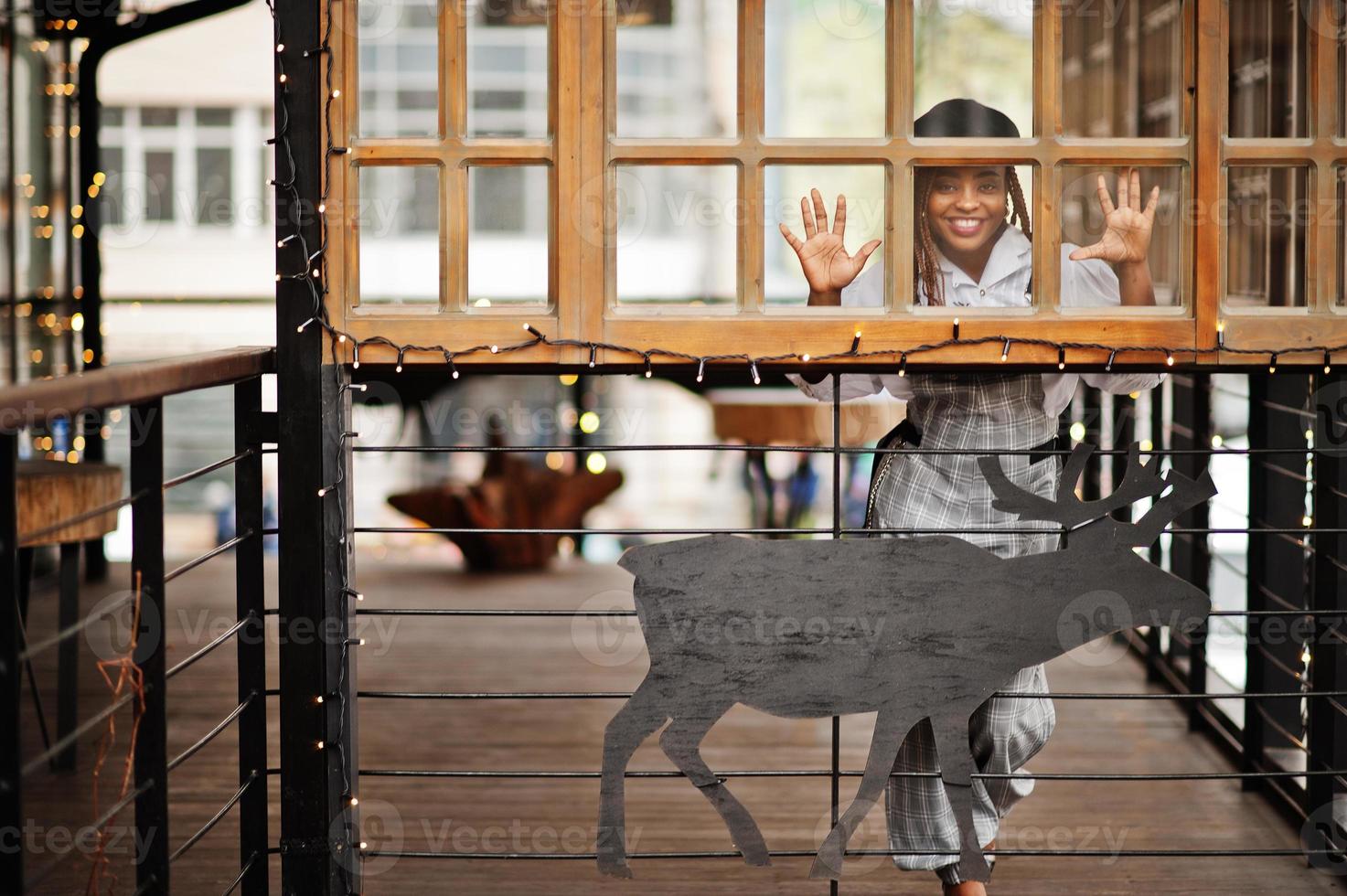 femme afro-américaine en salopette et béret posé sur une terrasse extérieure avec guirlande de décorations de noël et cerf. photo