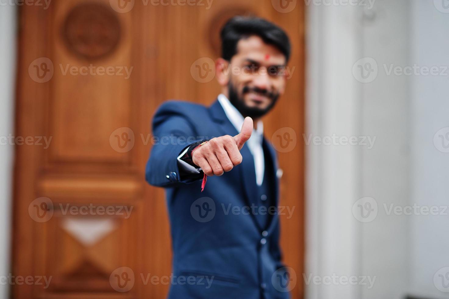 homme indien à la barbe élégante avec bindi sur le front, porter un costume bleu posé à l'extérieur contre la porte du bâtiment et montrer le pouce vers le haut. photo