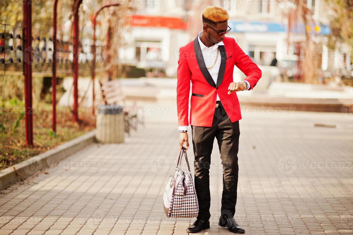 modèle d'homme afro-américain de mode en costume rouge, avec des cheveux en surbrillance et un sac à main posé dans la rue et regardant ses montres. photo