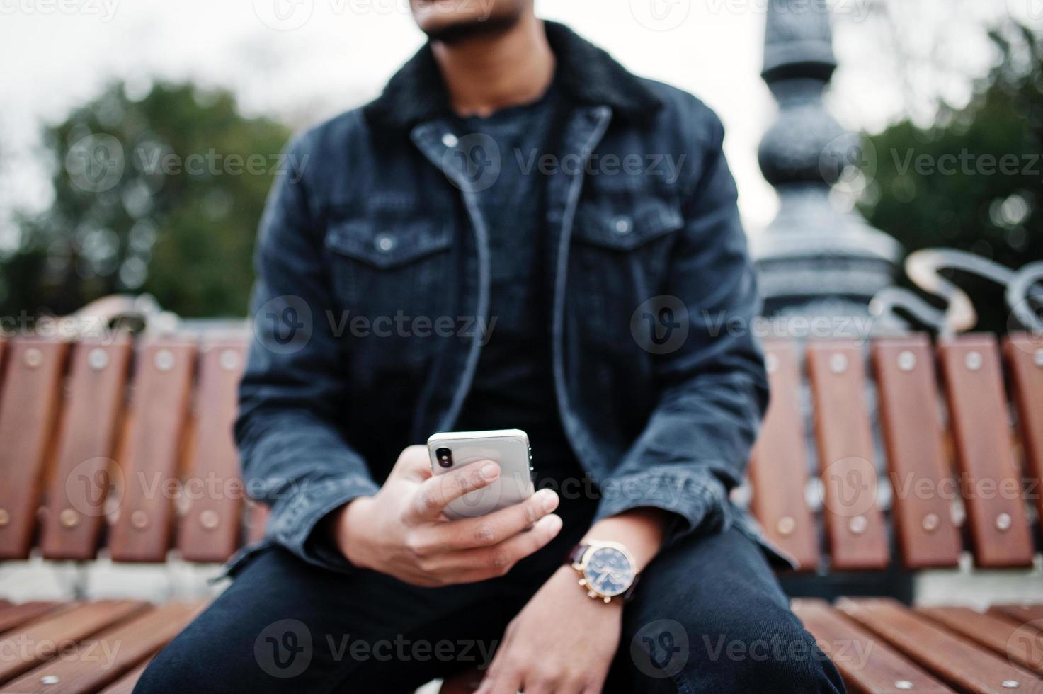 bel homme indien à la mode en veste de jeans noir posé à l'extérieur, assis sur un banc avec un téléphone portable à portée de main. photo