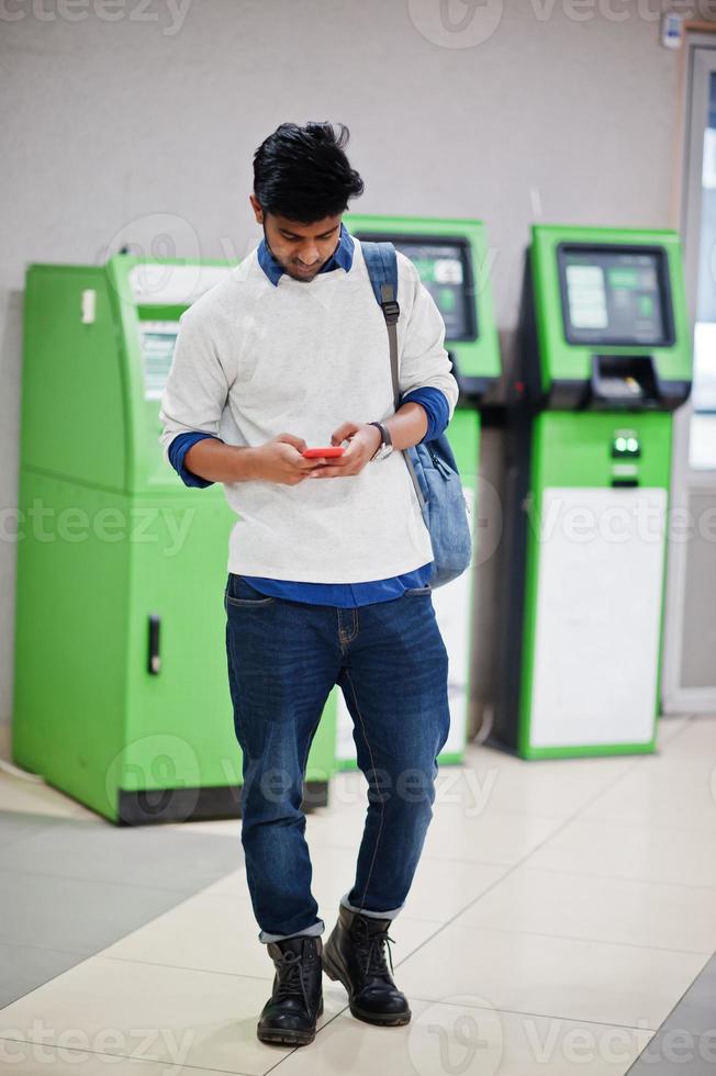 jeune homme asiatique élégant avec téléphone portable et sac à dos contre la rangée de guichets automatiques verts. photo