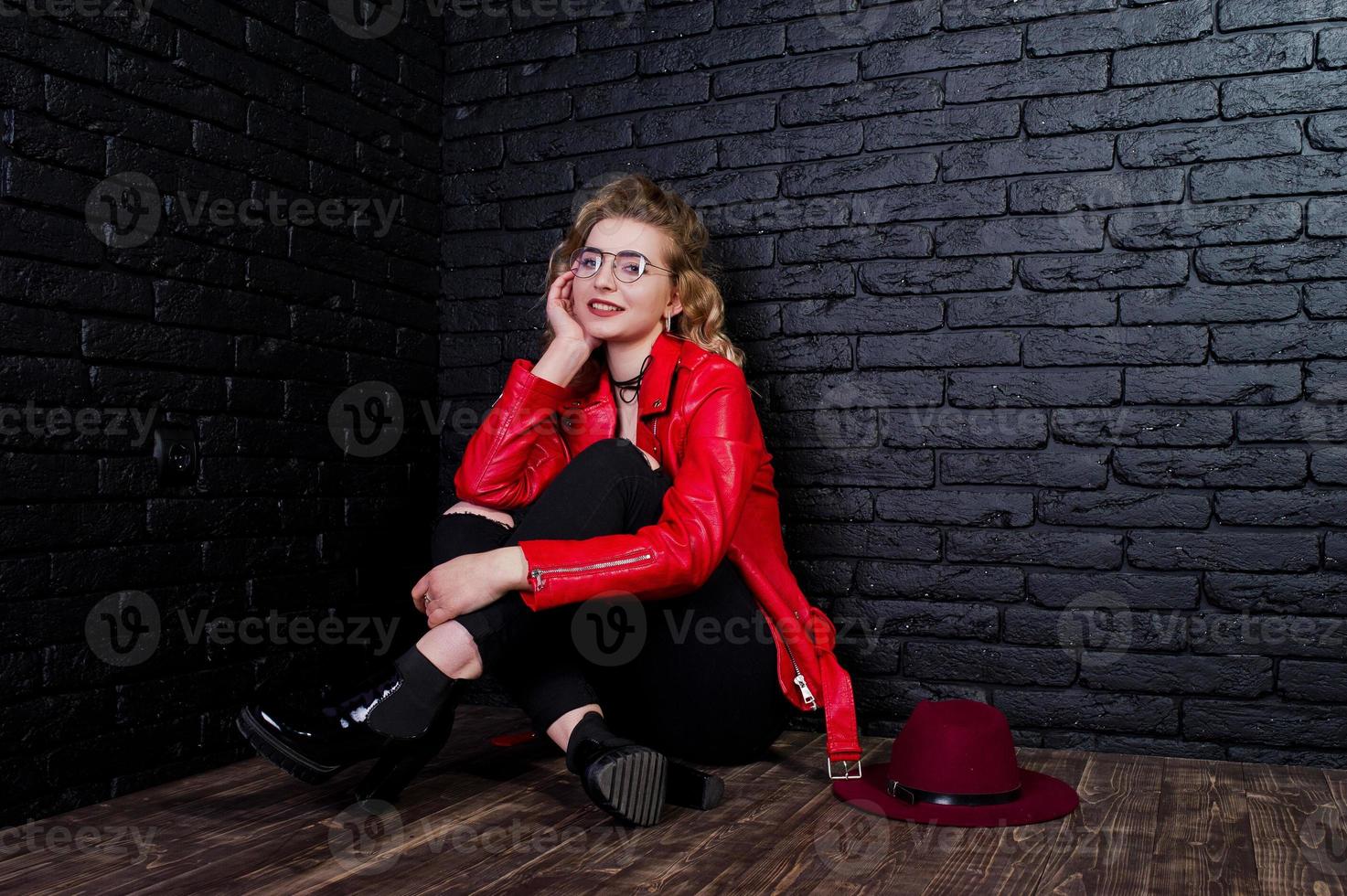 portrait en studio d'une fille blonde avec un chapeau rouge, des lunettes et une veste en cuir posées contre un mur de briques. photo