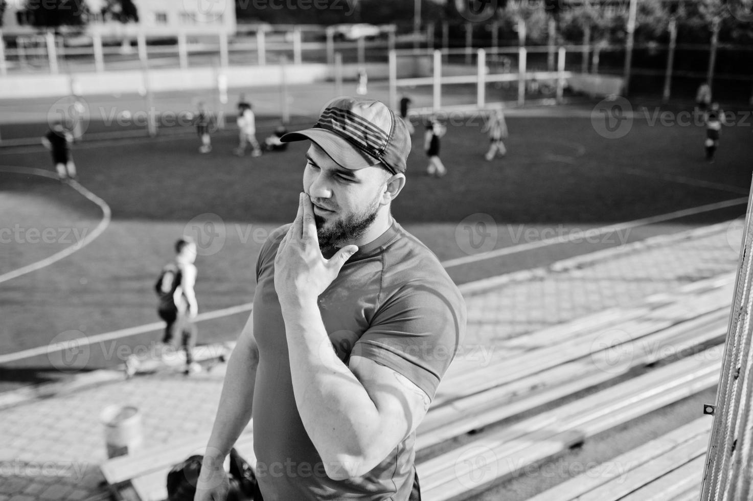 jeune homme musclé barbu brutal portant une chemise rouge, un short et une casquette au stade. photo