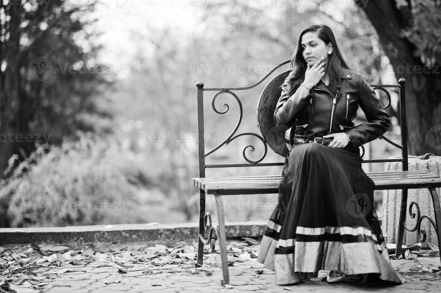 jolie fille indienne en robe saree noire et veste en cuir posée en plein air dans la rue d'automne et assise sur un banc. photo