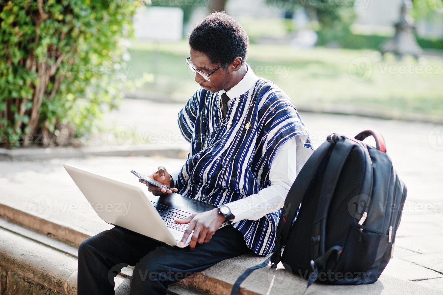 étudiant africain en vêtements traditionnels et lunettes avec sac à dos, travaillant sur un ordinateur portable avec un téléphone portable en plein air. photo