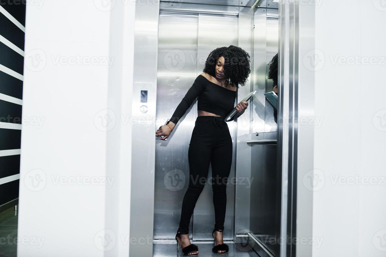 jeune belle femme d'affaires afro-américaine à la mode avec une coiffure afro en noir élégant, debout à l'ascenseur avec téléphone et ordinateur portable à portée de main. photo