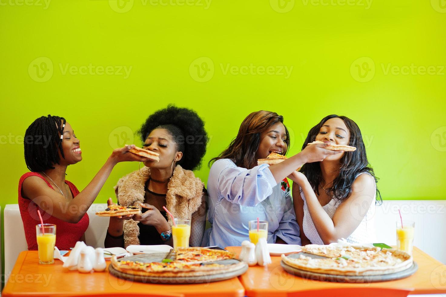 quatre jeunes filles africaines dans un restaurant aux couleurs vives mangeant des tranches de pizza dans les mains. photo
