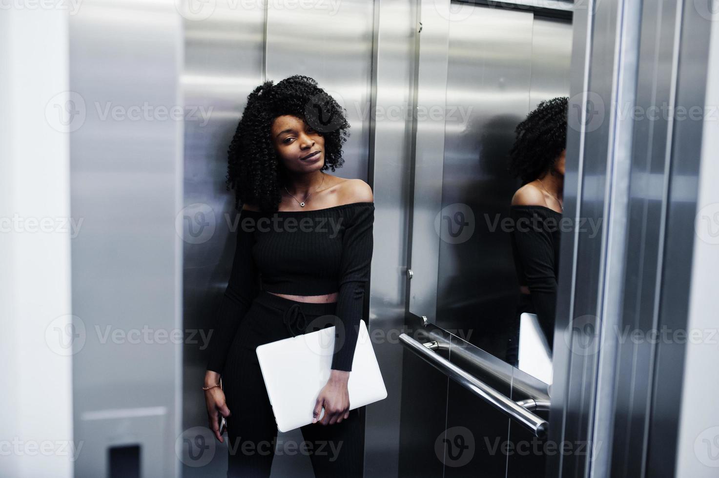 jeune belle femme d'affaires afro-américaine à la mode avec une coiffure afro en noir élégant, debout à l'ascenseur avec téléphone et ordinateur portable à portée de main. photo