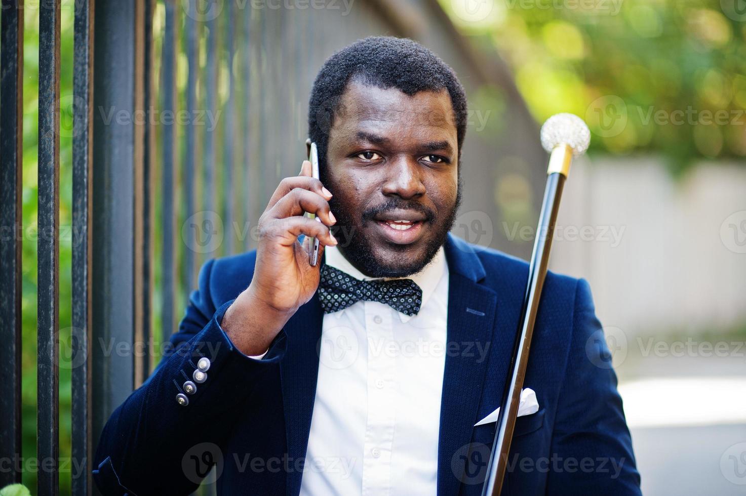 bel homme afro-américain à la mode en tenue de soirée et noeud papillon avec canne parlant au téléphone. photo