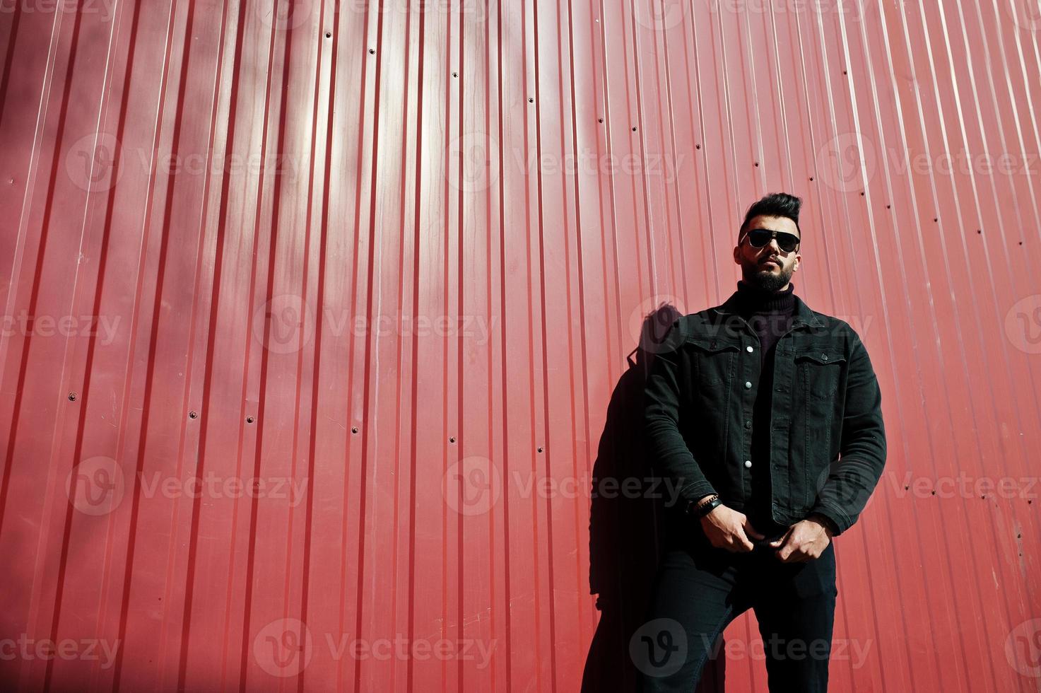 homme arabe de mode porter sur une veste en jean noir et des lunettes de soleil posées sur fond de mur en acier rouge. mec modèle arabe élégant et à la mode. photo