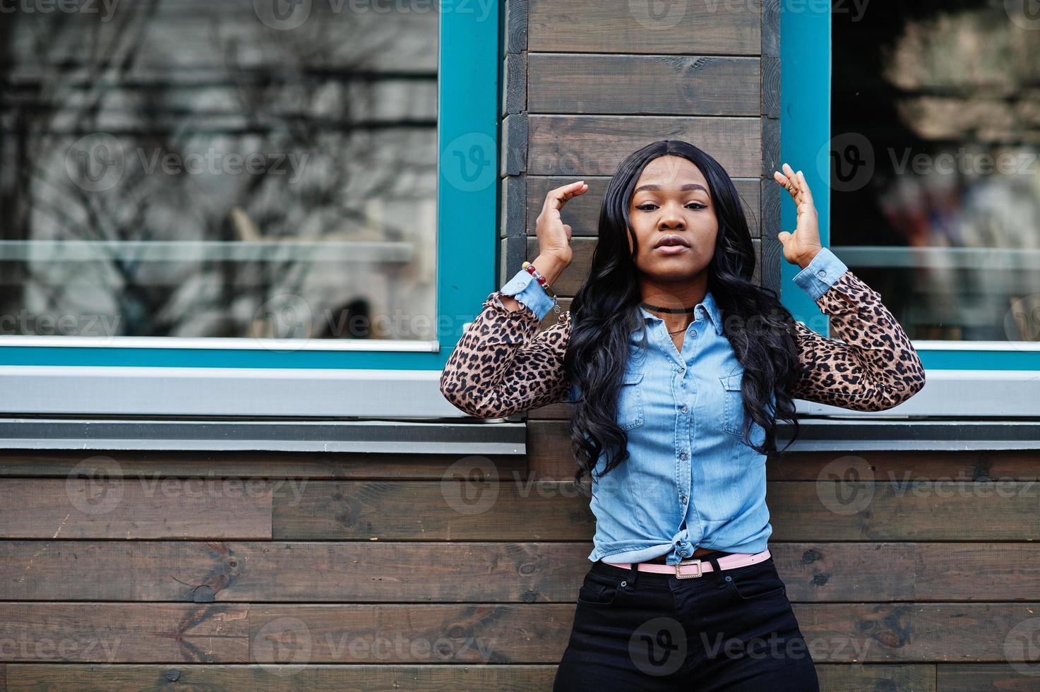 fille afro-américaine hipster portant une chemise en jean à manches léopard posant dans la rue contre une maison en bois avec des fenêtres. photo