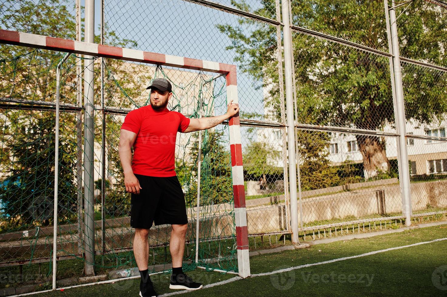 jeune homme musclé barbu brutal portant une chemise rouge, un short et une casquette au stade. photo