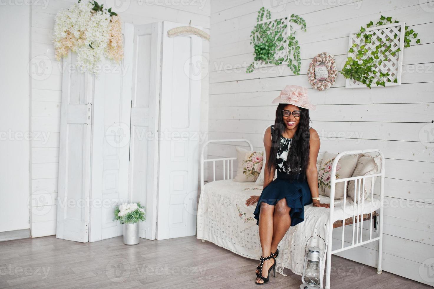 jolie femme afro-américaine à lunettes et chapeau posé dans la chambre assise sur un canapé vintage. photo