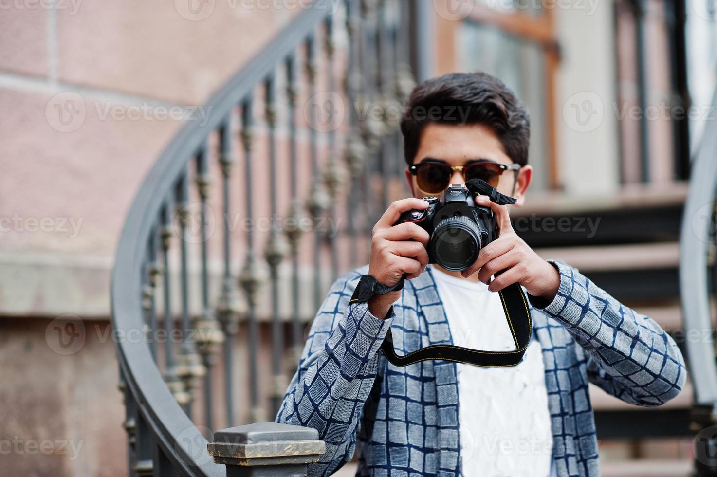 Un jeune photographe indien élégant à lunettes de soleil porte des lunettes de soleil décontractées posées en plein air contre des escaliers en fer avec un appareil photo reflex numérique à portée de main.