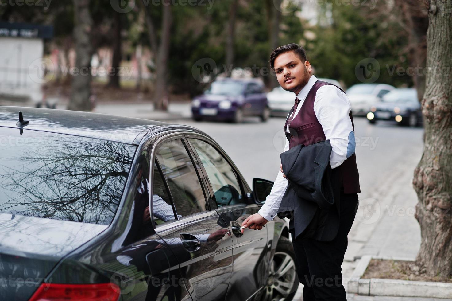 homme d'affaires indien élégant en tenue de soirée debout contre une voiture d'affaires noire dans la rue de la ville. ouvrir la porte de la voiture par clé. photo