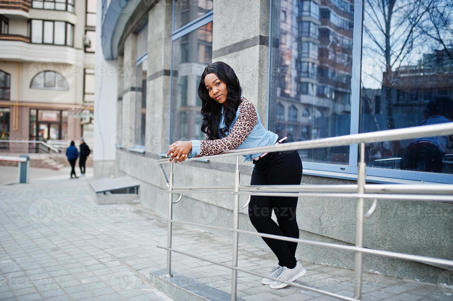 fille afro-américaine hipster portant une chemise en jean à manches léopard posant dans la rue contre un immeuble de bureaux moderne aux fenêtres bleues. photo