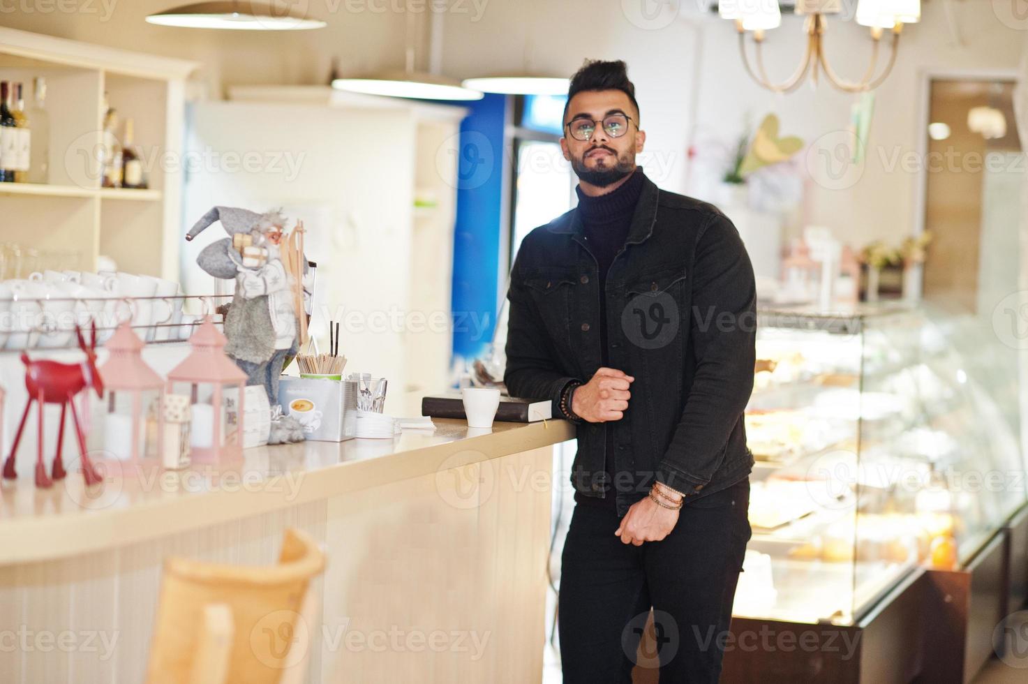 l'homme arabe porte une veste en jean noir et des lunettes au café boit du café au bar. mec modèle arabe élégant et à la mode. photo