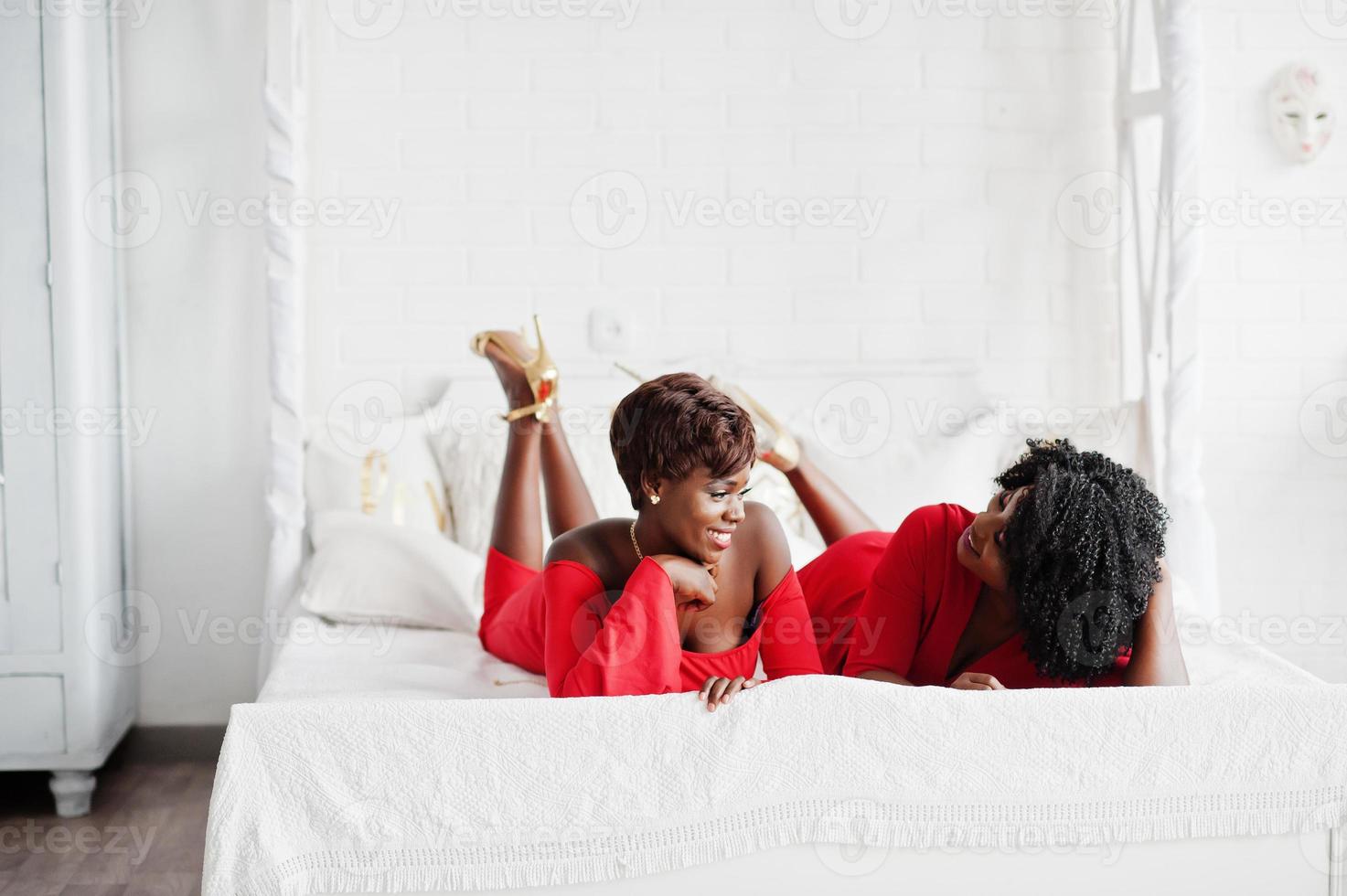 deux mannequins afro-américains de mode en robe de beauté rouge et talons hauts dorés, des femmes sexy posant une robe de soirée se trouvent sur le lit. photo