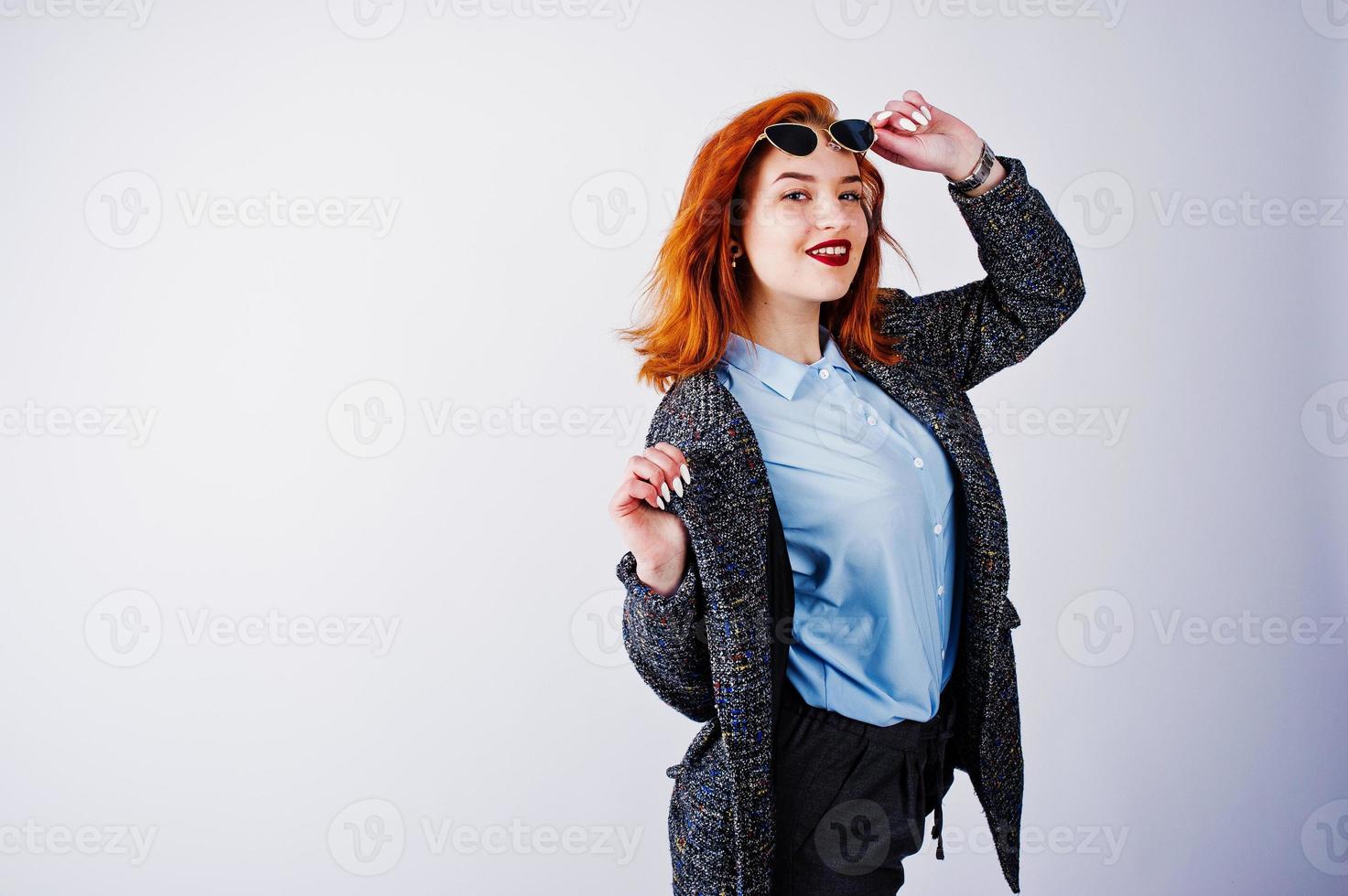 portrait d'une fantastique fille rousse en chemise bleue, pardessus gris posant avec des lunettes de soleil en studio. photo