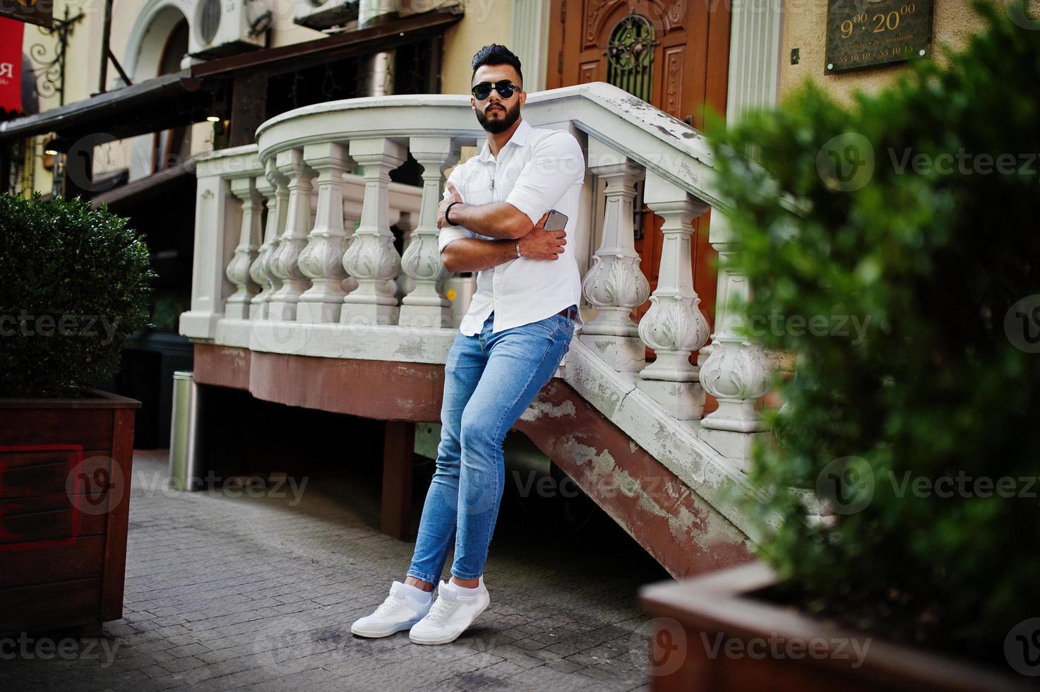 grand modèle d'homme arabe élégant en chemise blanche, jeans et lunettes de soleil posés dans la rue de la ville. Barbe mec arabe attrayant. photo