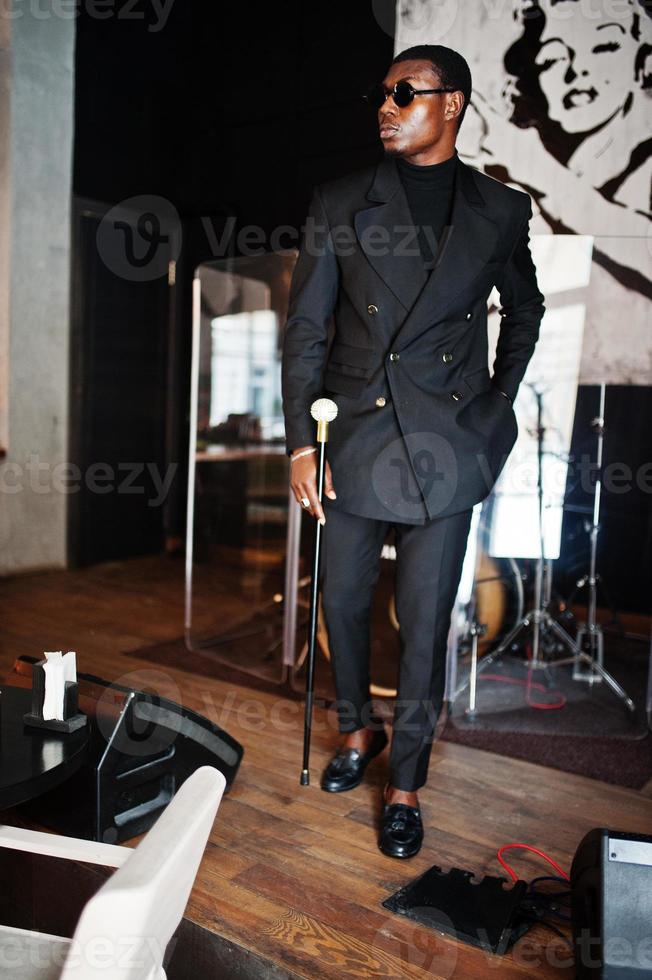 élégant gentleman afro-américain dans une élégante veste noire et des lunettes de soleil, tenant un bâton de marche rétro comme un flacon de canne ou une canne basculante avec une poignée en boule de diamant doré. riche homme afro à la mode. photo