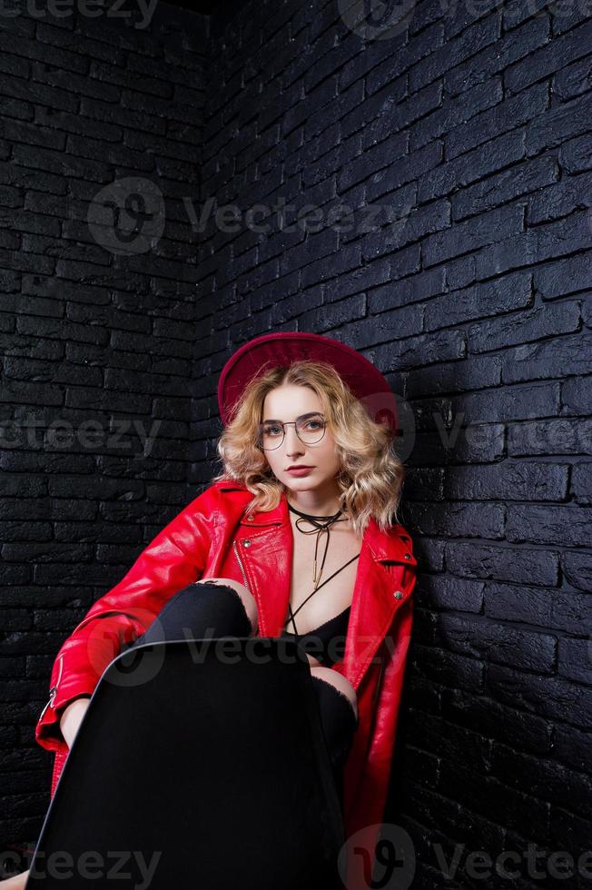 portrait en studio d'une fille blonde au chapeau rouge, lunettes et veste en cuir posée sur une chaise contre un mur de briques. photo