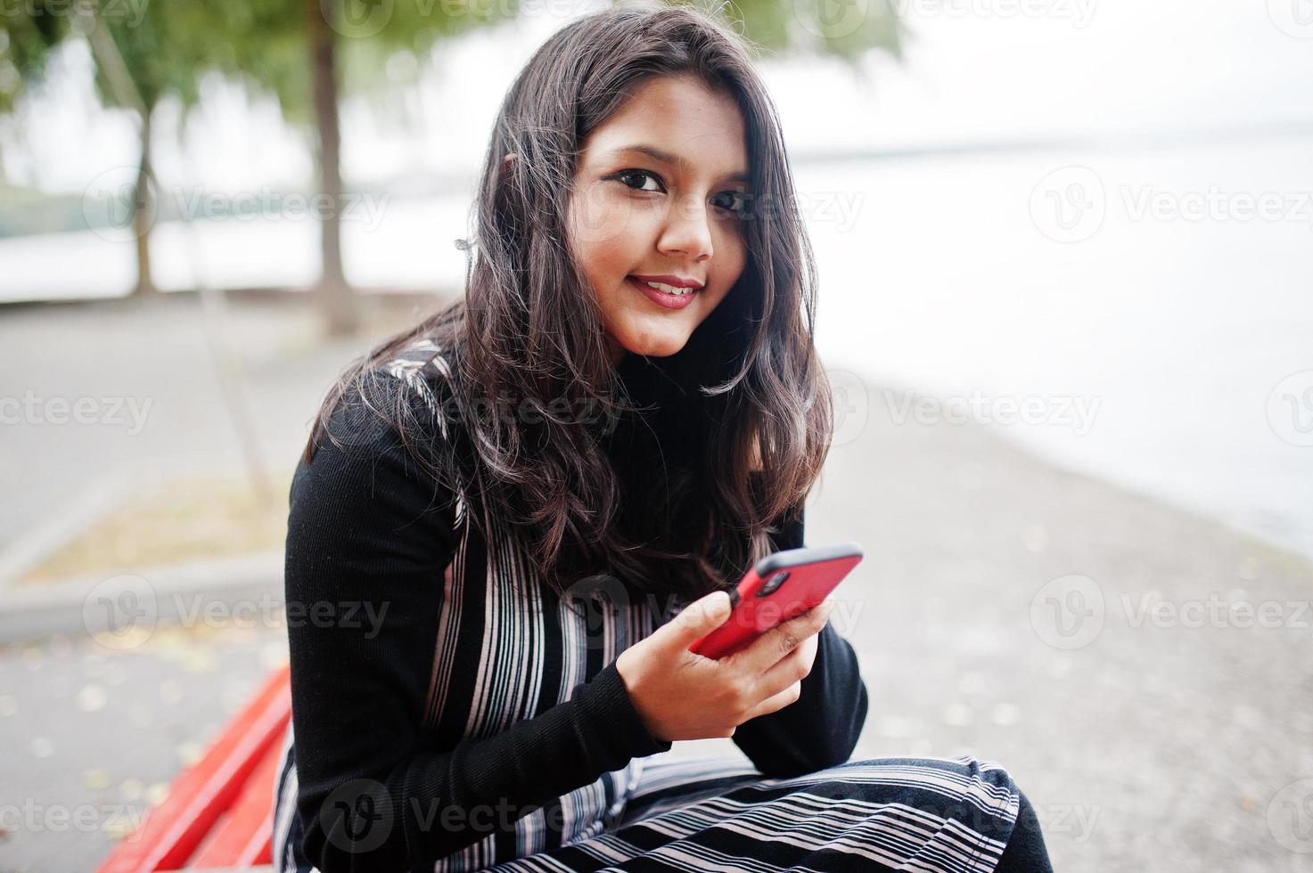 portrait d'une jeune belle adolescente indienne ou sud-asiatique en robe assise sur un banc avec un téléphone portable. photo