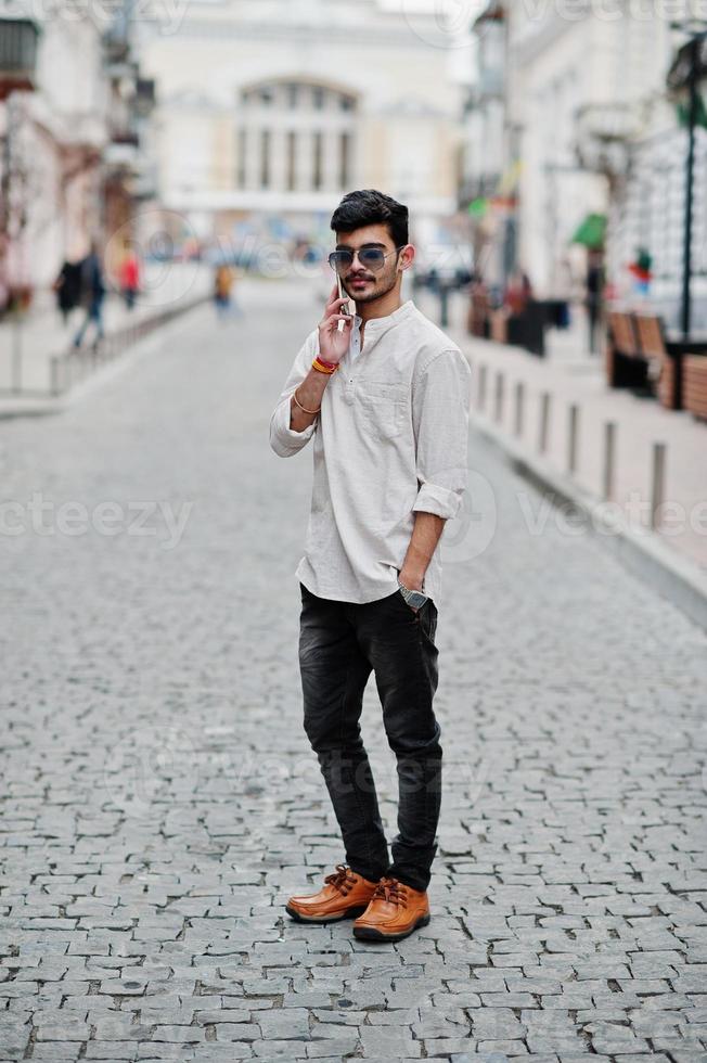 homme modèle indien élégant dans des vêtements décontractés et des lunettes de soleil posés à l'extérieur dans la rue de l'inde et parlant au téléphone mobile. photo