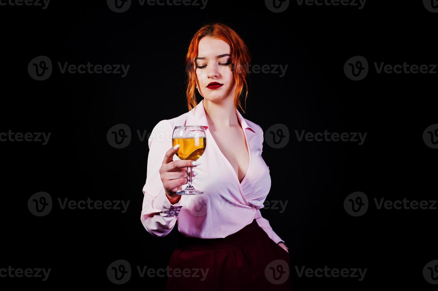 magnifique fille aux cheveux rouges en chemisier rose et jupe rouge avec un verre de vin à portée de main isolé sur noir. photo