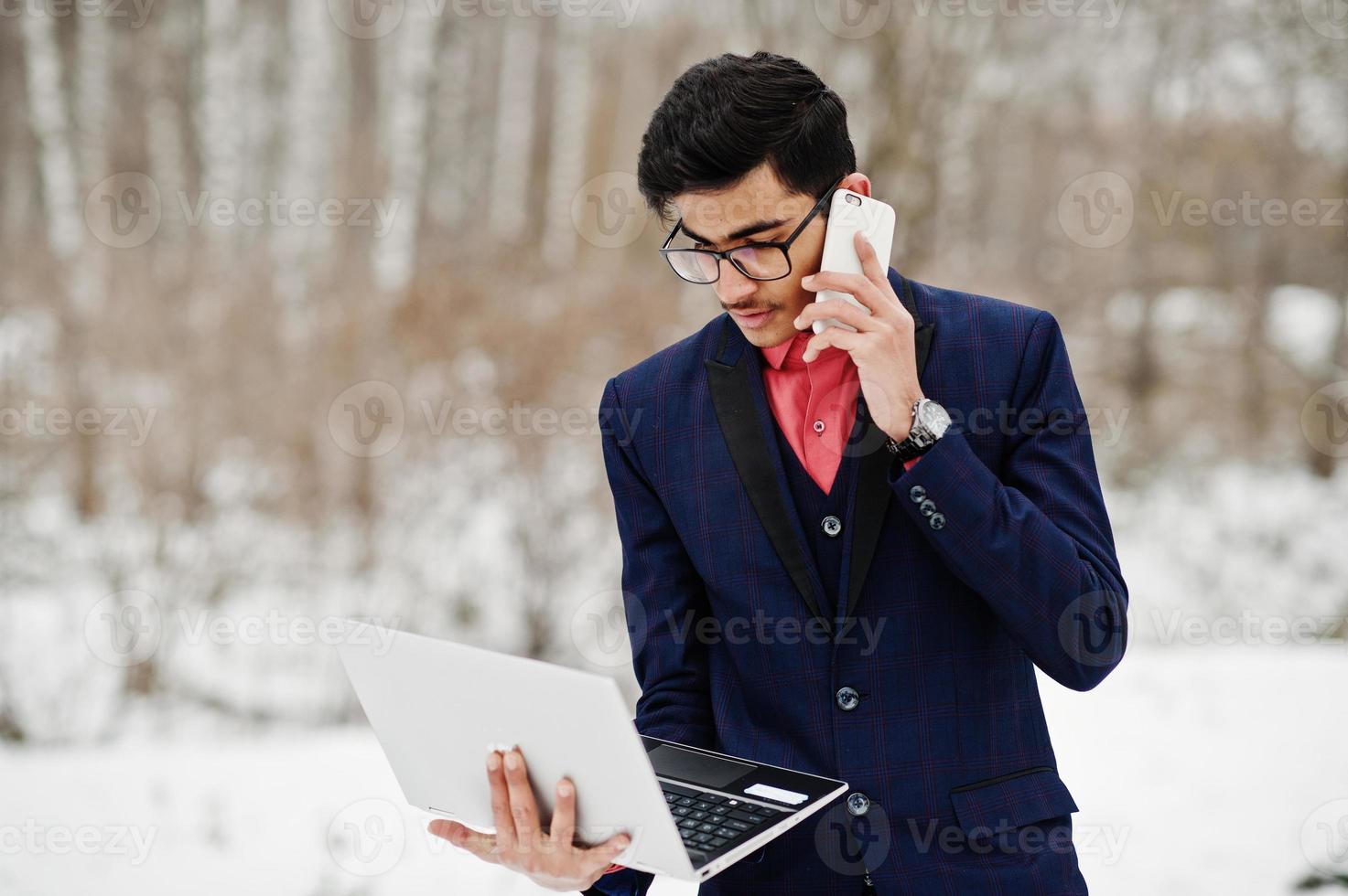 homme étudiant indien élégant en costume et lunettes posé à la journée d'hiver en plein air avec un ordinateur portable à la main, parlant au téléphone. photo