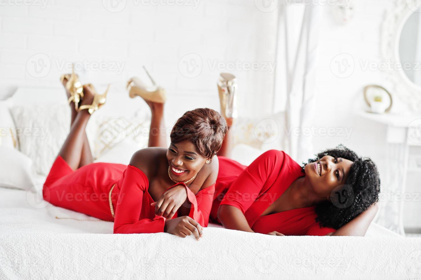 deux mannequins afro-américains de mode en robe de beauté rouge et talons hauts dorés, des femmes sexy posant une robe de soirée se trouvent sur le lit. photo