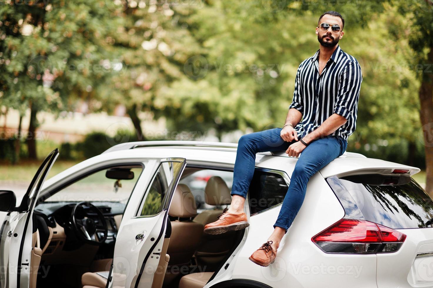un homme arabe réussi porte une chemise rayée et des lunettes de soleil assis sur le toit de sa voiture suv blanche. photo