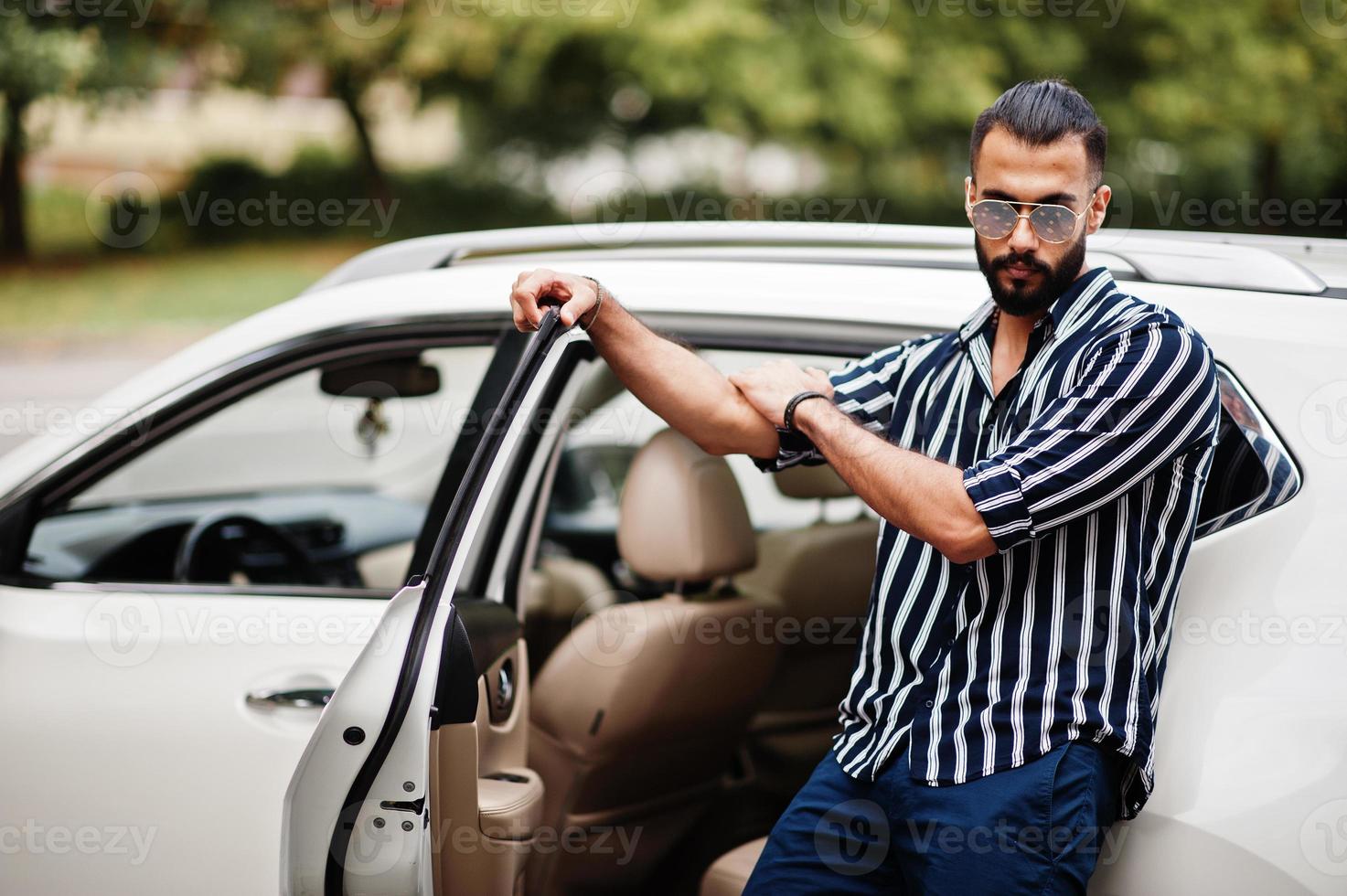 un homme arabe réussi porte une chemise rayée et des lunettes de soleil pose près de sa voiture suv blanche. hommes arabes élégants dans les transports. photo