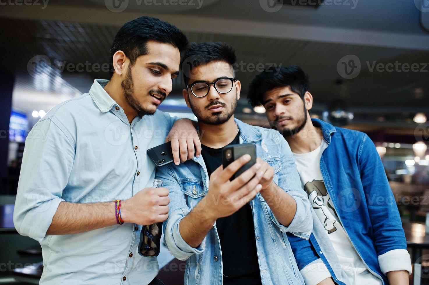 trois hommes asiatiques élégants portent des jeans en regardant les téléphones portables et boivent du soda contre la table du club. photo