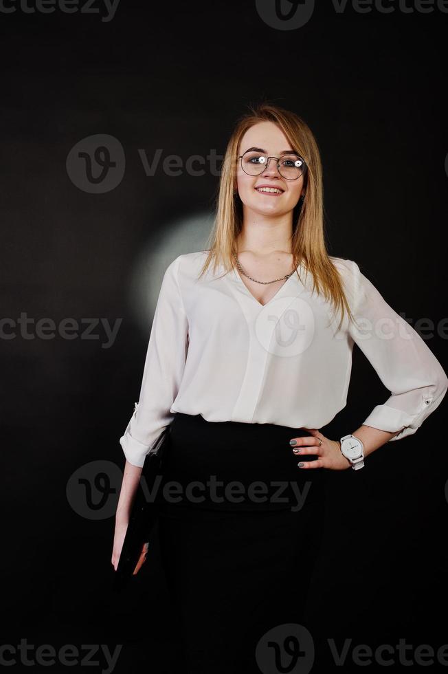 portrait en studio d'une femme d'affaires blonde à lunettes, chemisier blanc et jupe noire tenant un ordinateur portable sur fond sombre. femme réussie et concept de fille élégante. photo