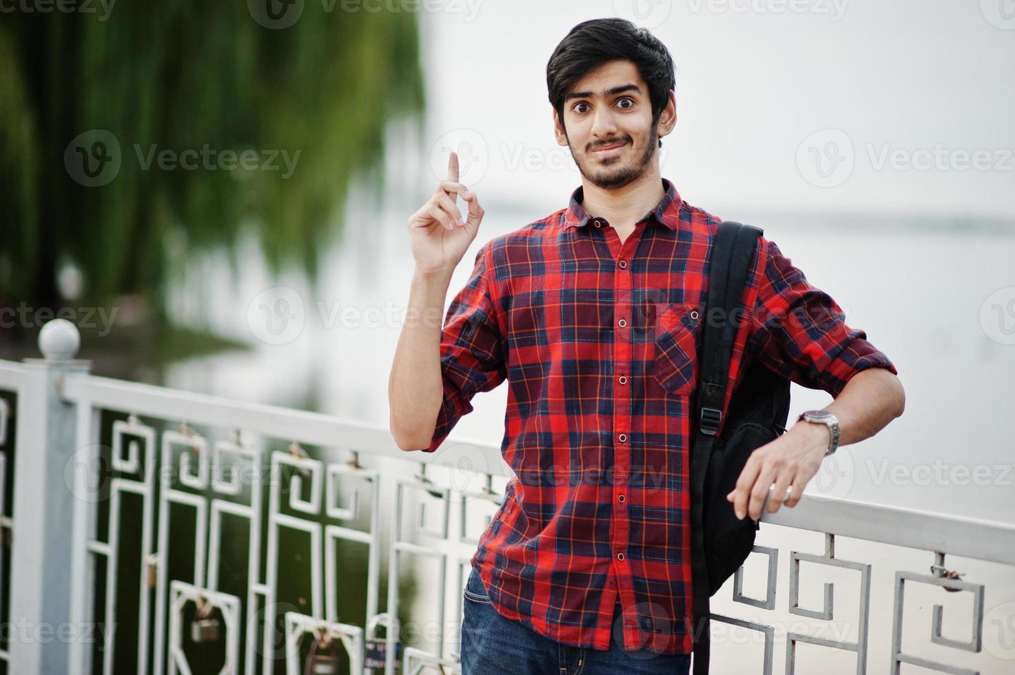 jeune étudiant indien en chemise à carreaux et jeans avec sac à dos posé sur la ville du soir contre le lac, montre une émotion surprise. photo