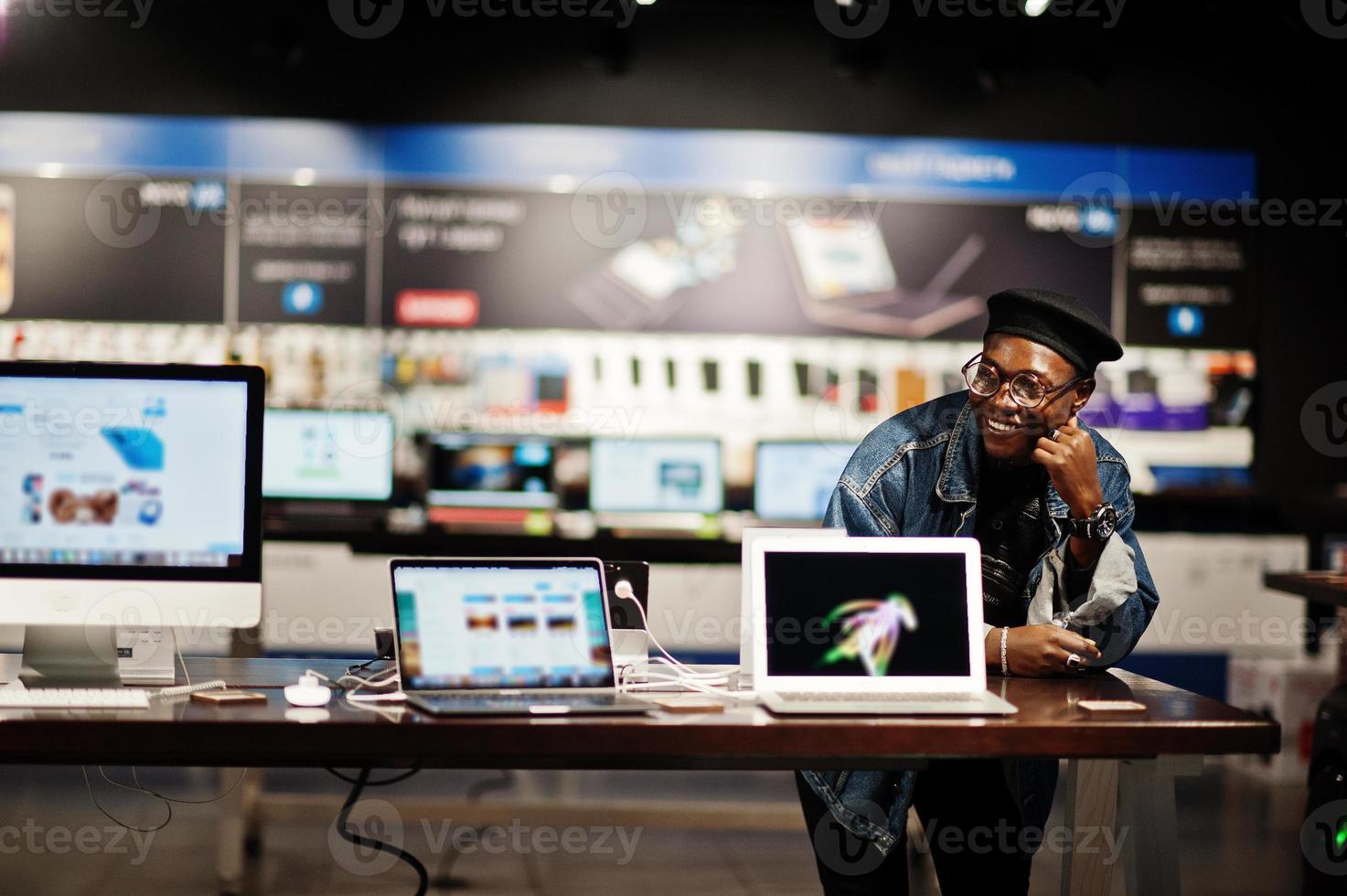 homme afro-américain décontracté élégant à la veste en jean et au béret noir contre le nouvel écran d'ordinateur portable au magasin d'électronique. photo