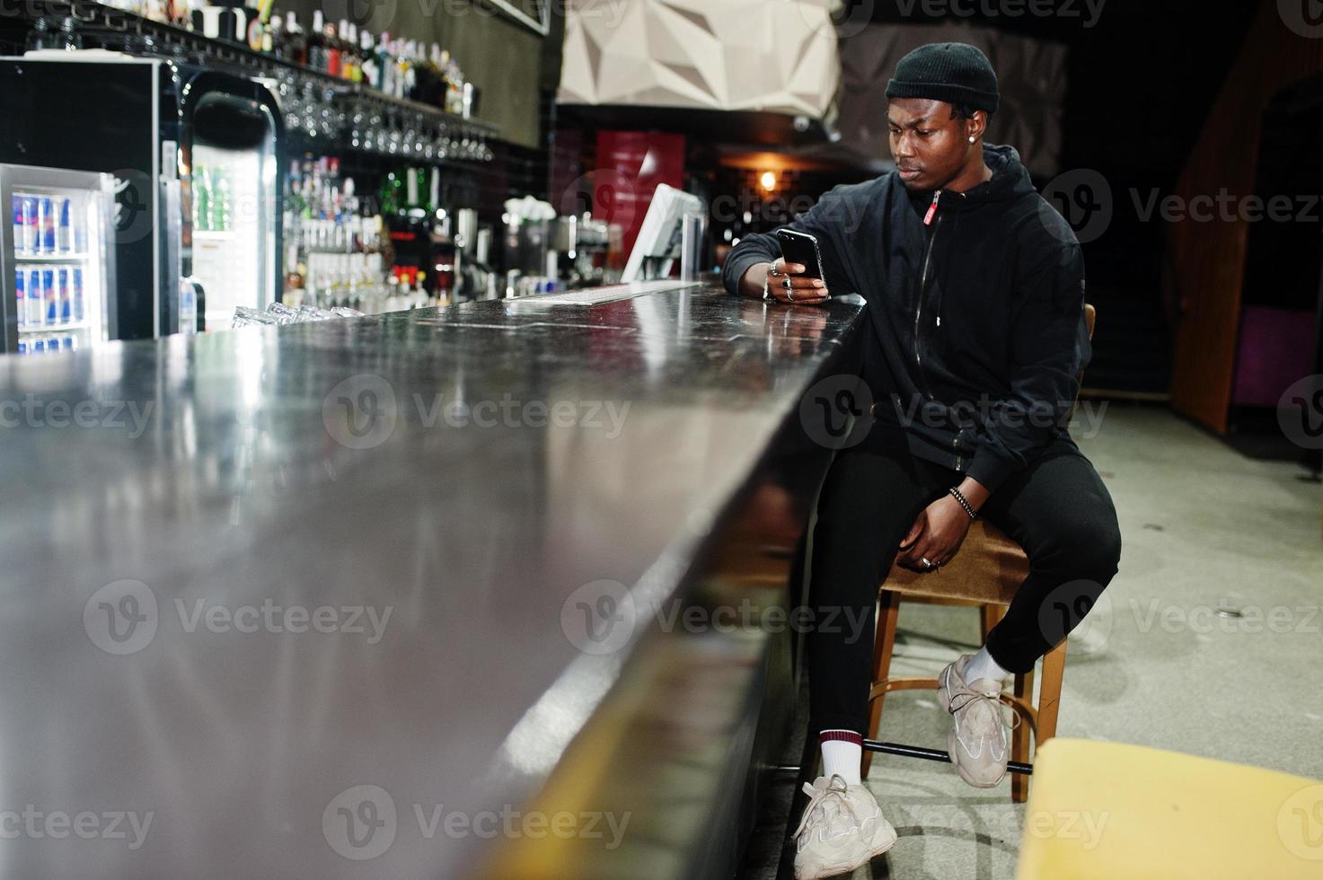 bel homme afro-américain posant à l'intérieur de la boîte de nuit en chapeau noir, assis sur le comptoir du bar. photo