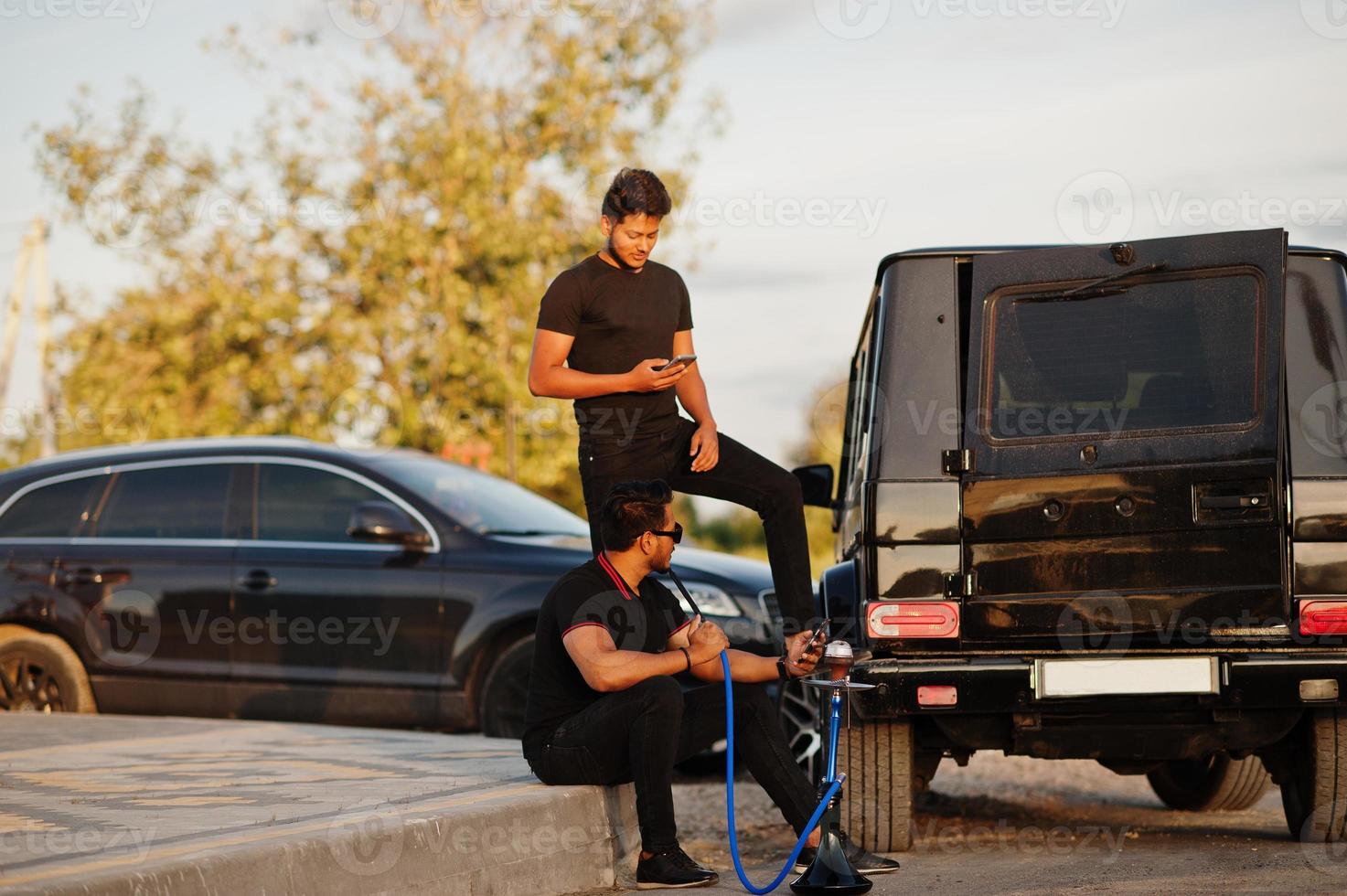 deux frères asiatiques portent un homme tout noir posé près de voitures suv et fument le narguilé. photo