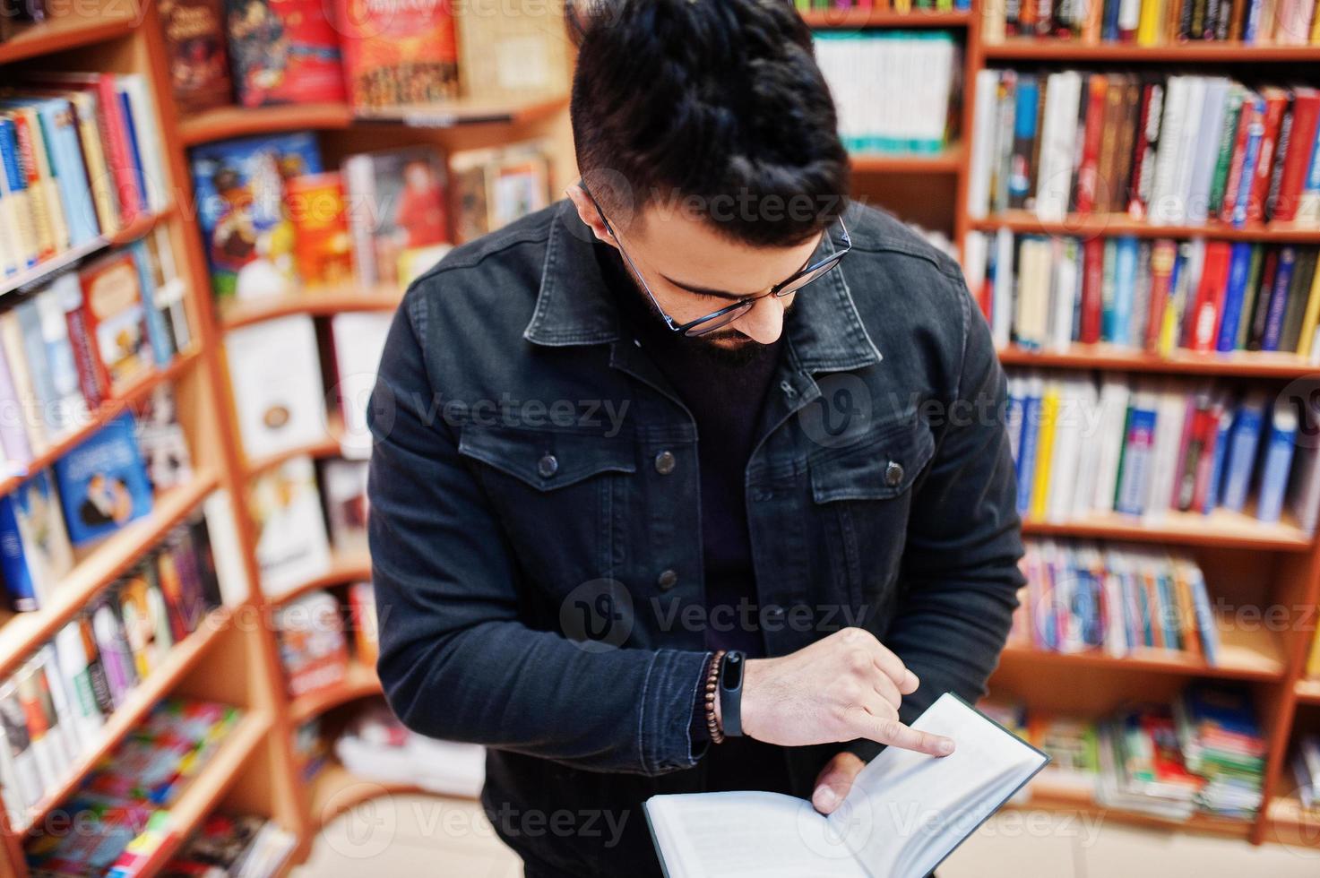 grand étudiant arabe intelligent, portant une veste en jean noir et des lunettes, à la bibliothèque avec un livre à portée de main. photo