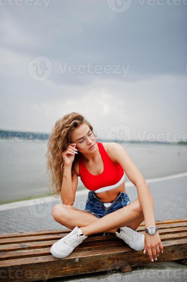 fille modèle bouclée sexy en haut rouge, short en jean jeans, lunettes et baskets posées sur un banc contre le lac. photo