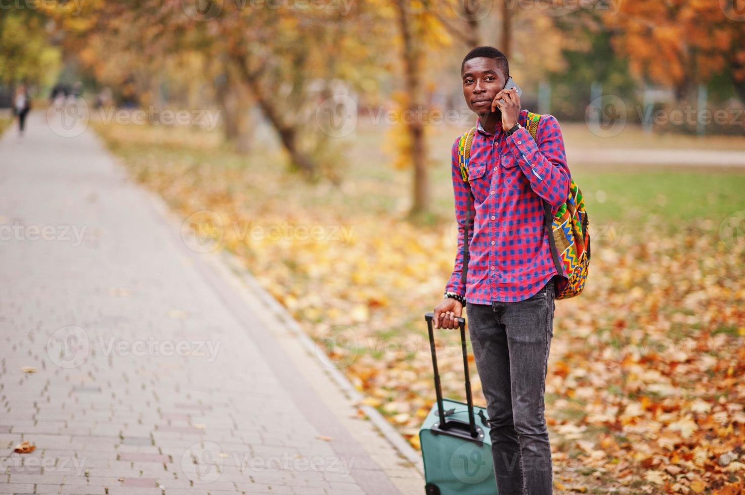 homme afro-américain en chemise à carreaux, avec valise et sac à dos.  voyageur d'homme noir au parc d'automne parlant au téléphone portable.  10570233 Photo de stock chez Vecteezy