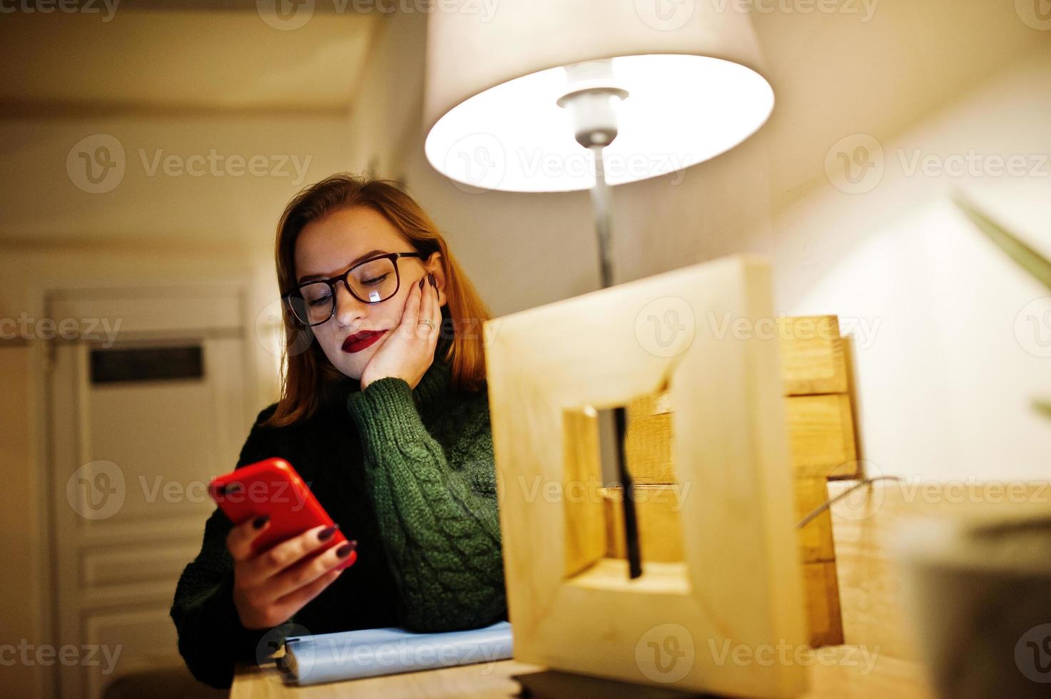 joyeuse jeune belle femme rousse à lunettes, portant un pull en laine chaude verte, assise au café et utilisant son téléphone portable. photo