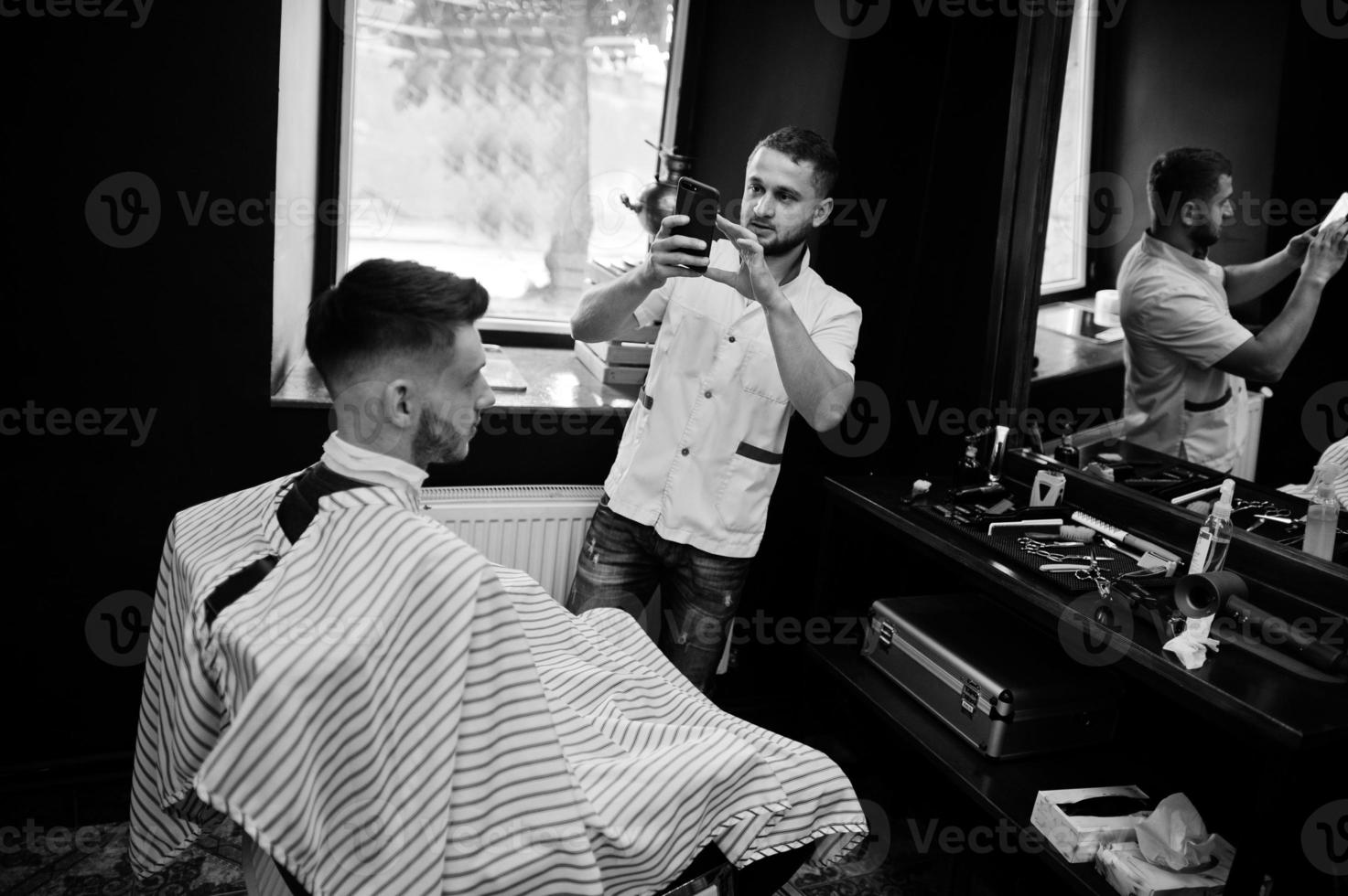 jeune homme barbu se coupe les cheveux par un coiffeur assis sur une chaise au salon de coiffure. âme de barbier. maître faisant la photo au téléphone.